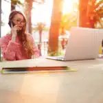 Charming woman having talking conversation on mobile phone while sitting at the table in sidewalk cafe with net-book, smart female speaking on cell telephone while rest after work on laptop computer contemplating $60,000 a Year Is How Much an Hour?