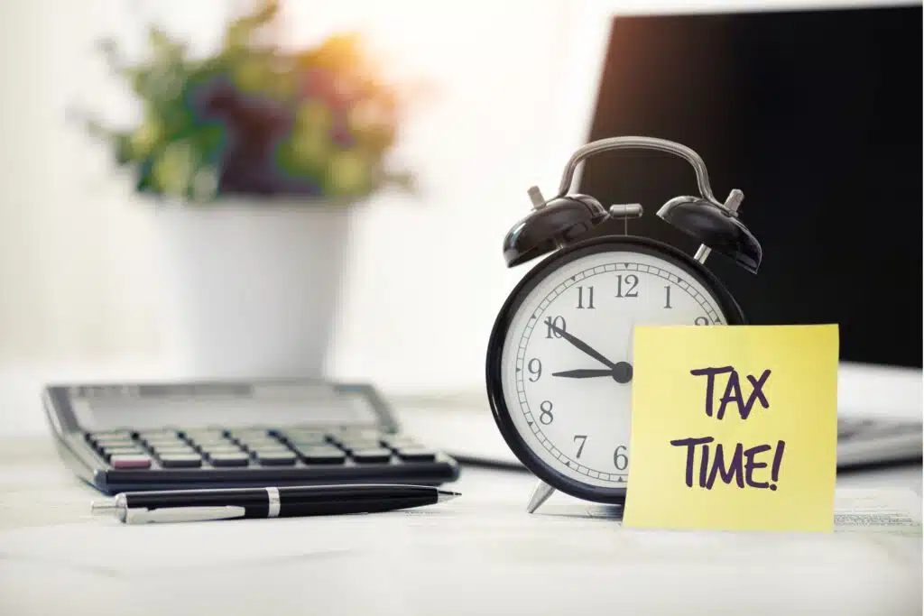 a sticky note with the words tax time leaning against an alarm clock on a desktop