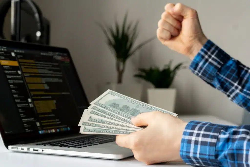 a man pumps his fist while sitting in front of a laptop holding a wad of cash in his left hand. 